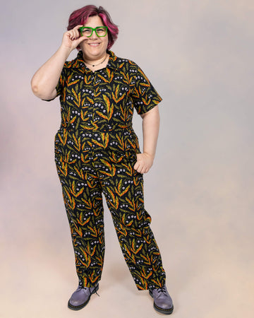 Jackie Brand smiles confidently while wearing a short-sleeve Run & Fly jumpsuit. The jumpsuit showcases a bold Halloween-inspired print by Clare Lewis, paired with purple boots and green glasses.