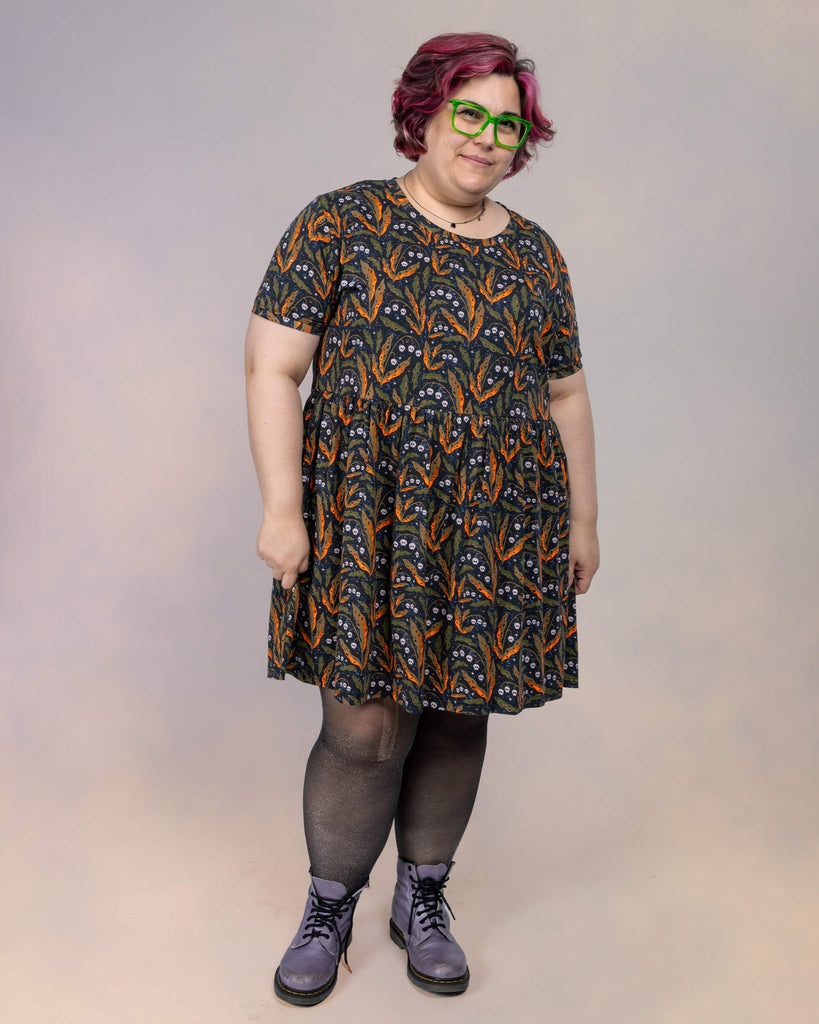 Jackie Brand wears a Run & Fly smock dress designed by Clare Lewis. The dress features a whimsical autumnal print with orange and green leaves, tiny skull motifs, and stars, paired with sheer black tights and purple boots.