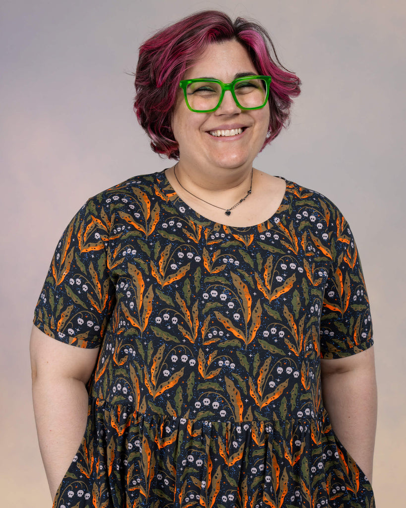 Close-up of Jackie Brand smiling while wearing the Run & Fly smock dress. The Clare Lewis-designed fabric showcases orange and green leaves intertwined with white skull details on a black background.