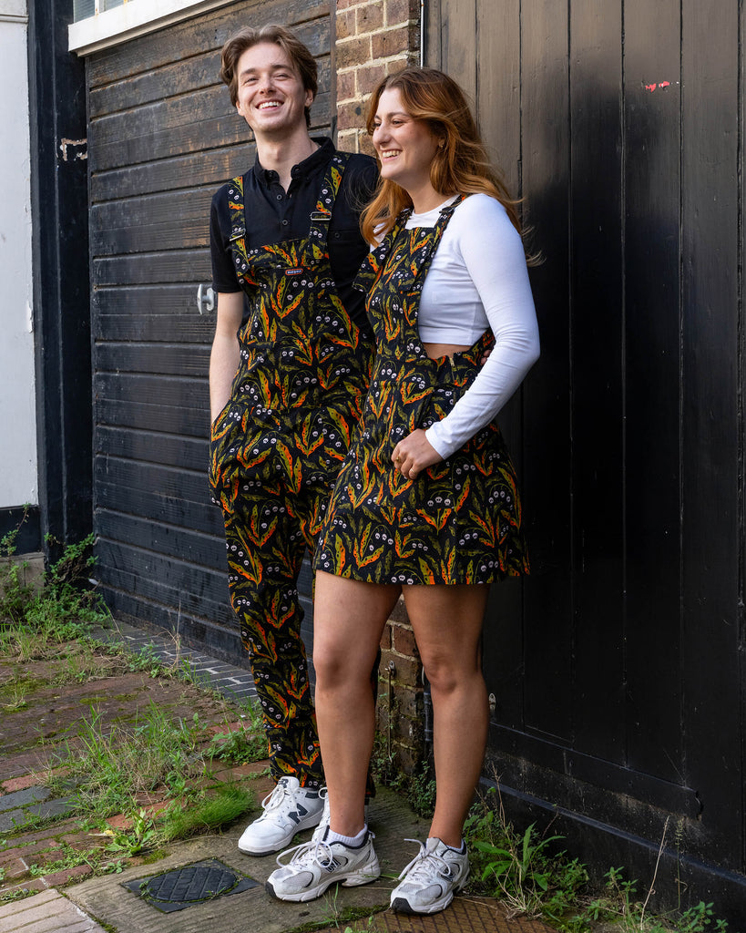 Two models smiling in matching Run & Fly outfits, featuring a bold skull and leaf print. The male model wears dungarees, and the female model wears a pinafore dress. Run & Fly is a small British brand known for its ethical, slow fashion and quirky designs. These coordinating looks are perfect for those who want to stand out while supporting sustainable, alternative fashion.