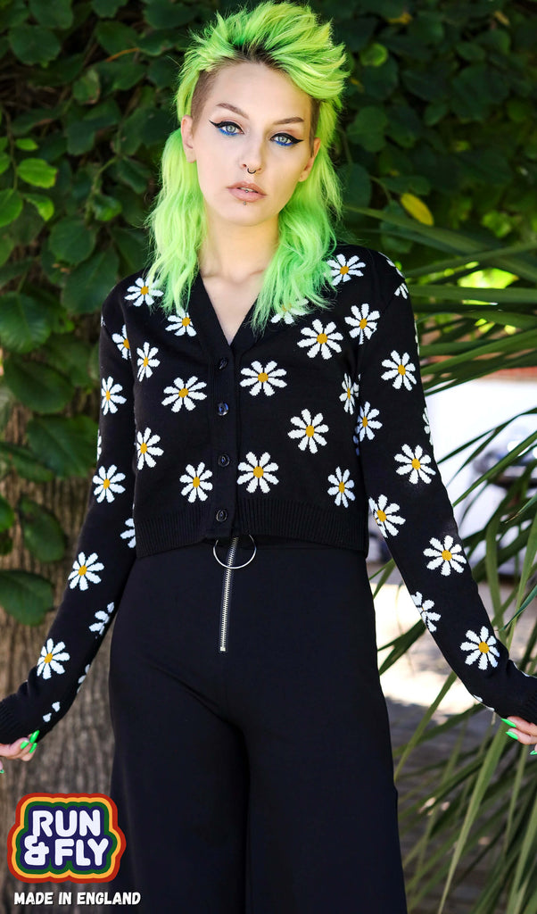 Green haired model is stood nearby a tree wearing the daisy chain black cropped cardigan and black trousers. They are facing the camera with both hands out by their sides. Photo has been cropped from the knees up.