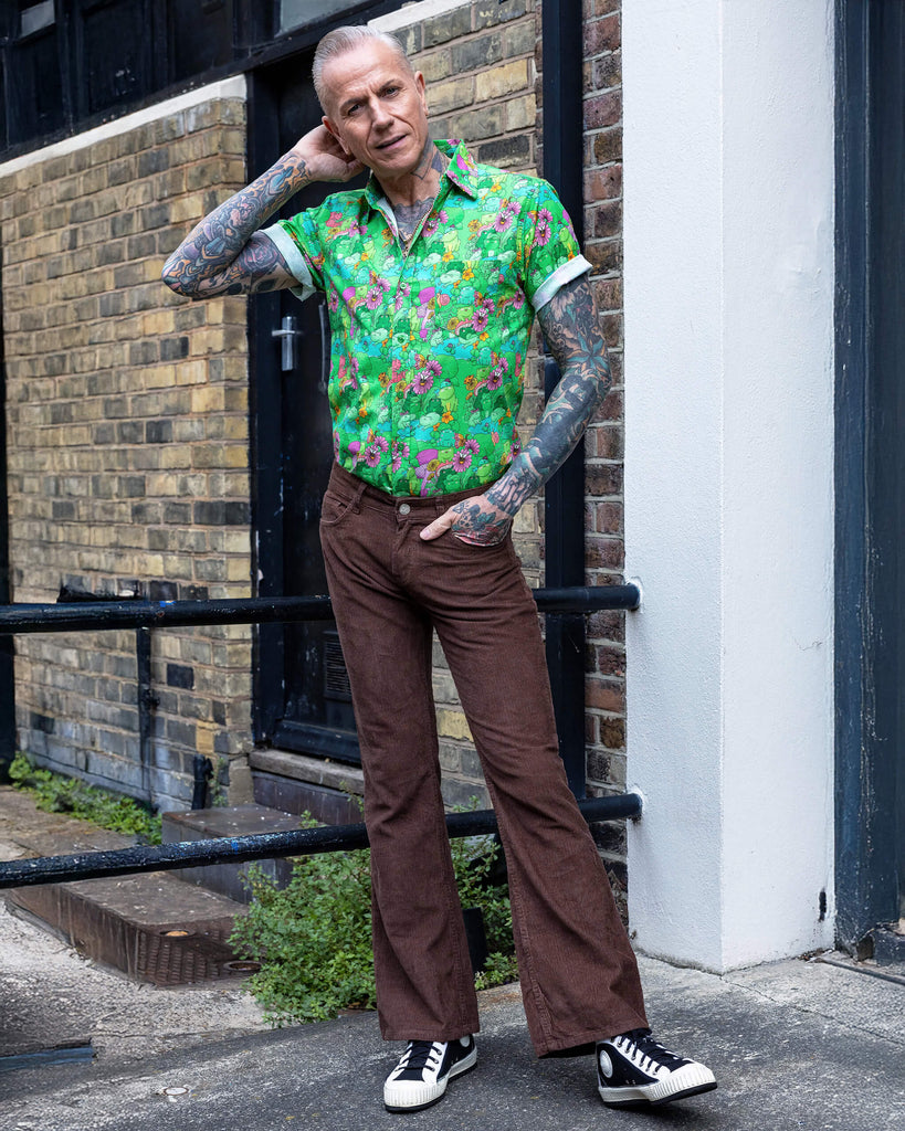 James a tattooed white male with silver hair in his 50's is posing wearing a frog print shirt and brown corduroy flares stood in a mews in Hove UK