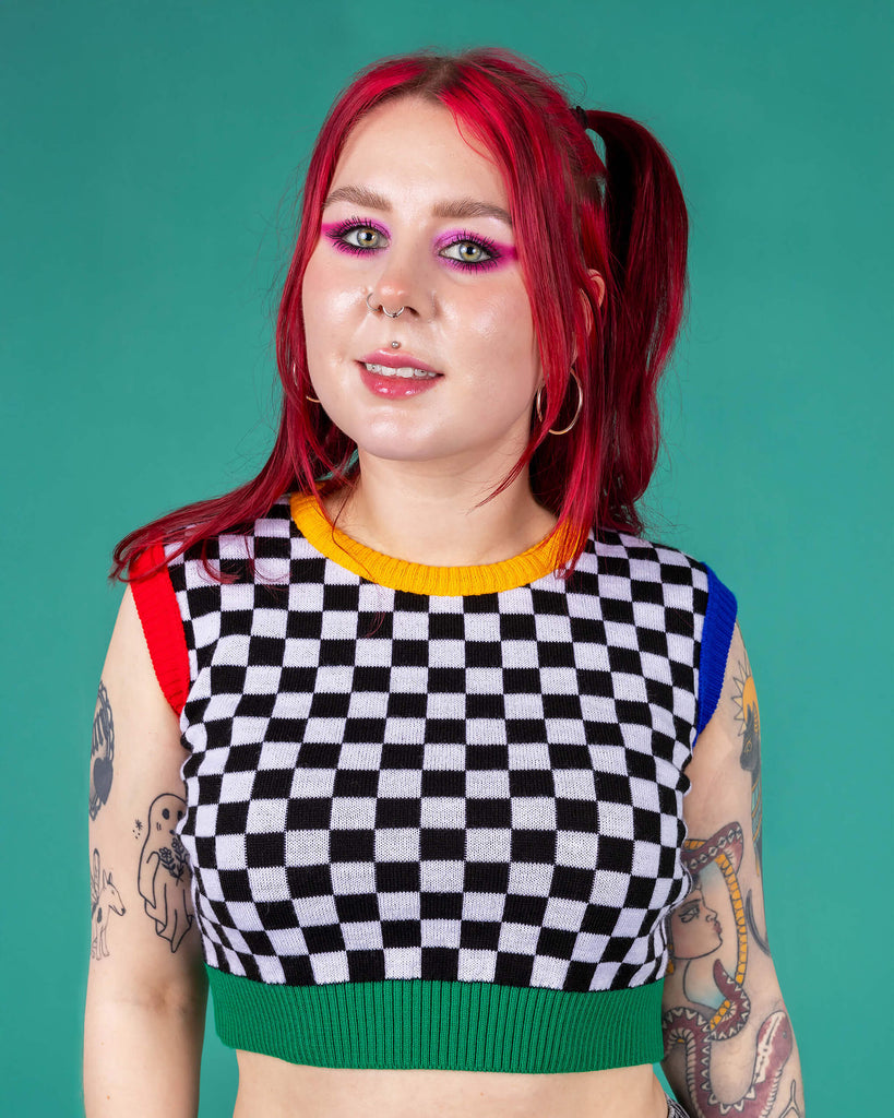 Flo wearing the Run and Fly Colour Block Checkerboard Cropped Knitted Tank Top on a teal background. She has styled the all over black and white checkerboard vest with a yellow trim neck, red left arm trim, blue right arm trim and green hem trim with oversized y2k cargo trousers, silver hoop earrings and neon pink eye makeup. Flo has red hair in bunches with piercings and tattoos. She is facing forward smiling with both hands resting by her side.