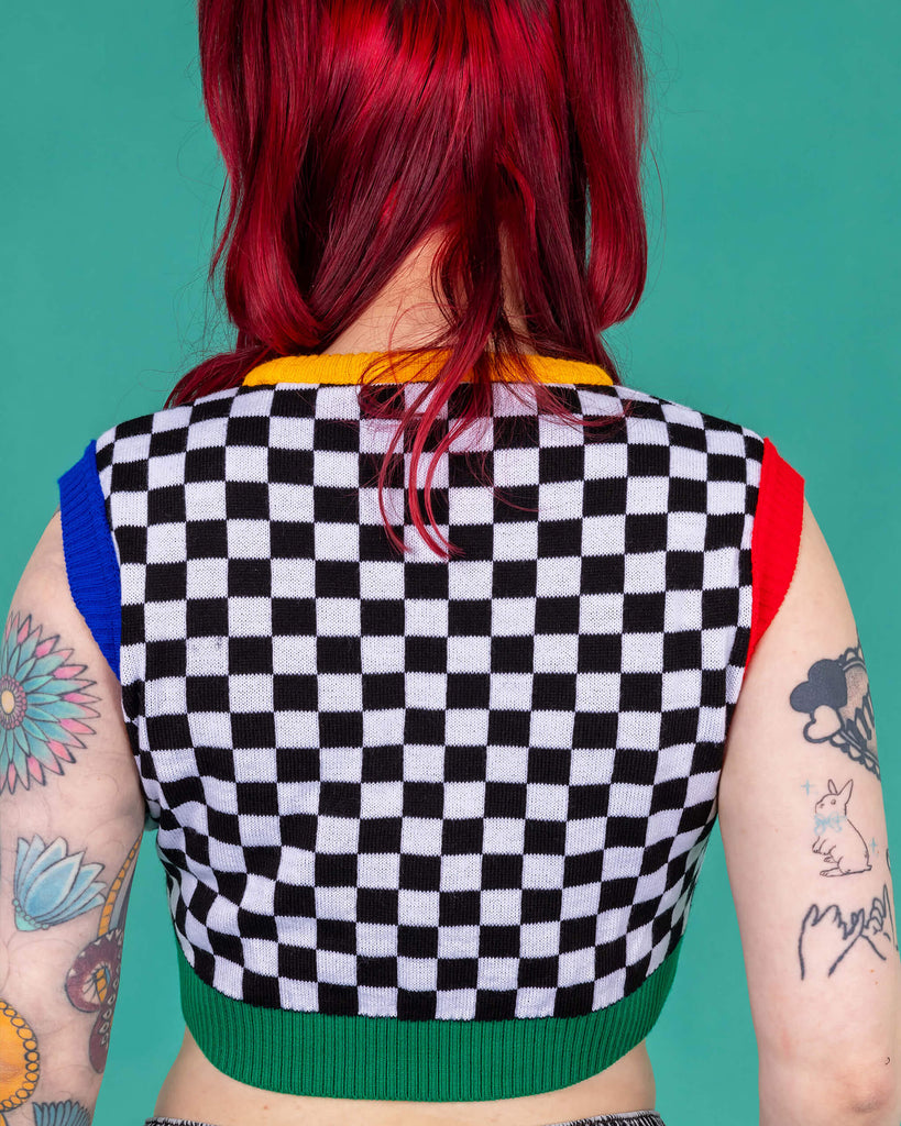 Flo wearing the Run and Fly Colour Block Checkerboard Cropped Knitted Tank Top on a teal background. She has styled the all over black and white checkerboard vest with a yellow trim neck, red left arm trim, blue right arm trim and green hem trim with oversized y2k cargo trousers, silver hoop earrings and neon pink eye makeup. Flo has red hair in bunches with piercings and tattoos. She is facing away from camera with both hands resting by her side.