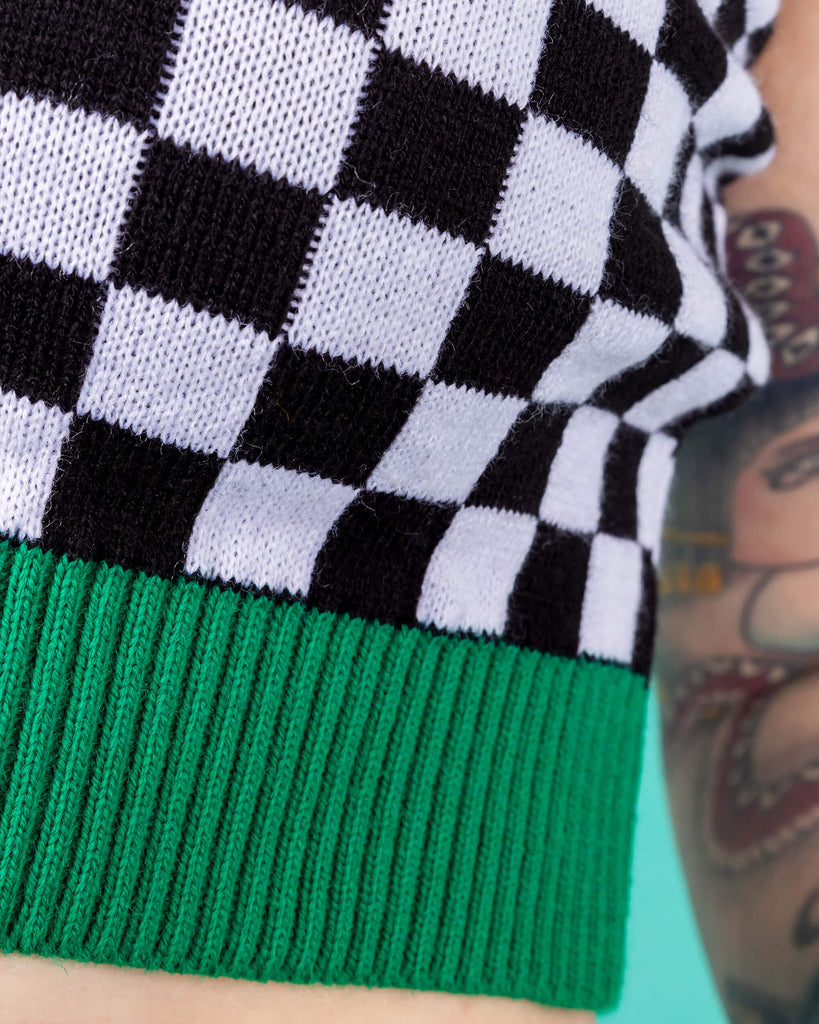 Flo wearing the Run and Fly Colour Block Checkerboard Cropped Knitted Tank Top on a teal background. She has styled the all over black and white checkerboard vest with a yellow trim neck, red left arm trim, blue right arm trim and green hem trim with oversized y2k cargo trousers, silver hoop earrings and neon pink eye makeup. Flo has red hair in bunches with piercings and tattoos. Close up of the lower green hem and checkerboard middle of flo facing forward with her arm resting by her side.