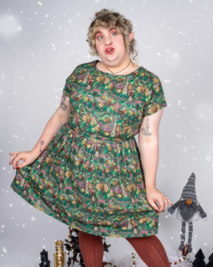 Maddie wears a festive dress with a print of cosy cottages and forest scenes, paired with earthy brown tights. Maddie stands playfully, leaning slightly, with a quirky expression, surrounded by a wintery setup with snowflakes falling, small black Christmas trees, and nutcracker figurines in the background.