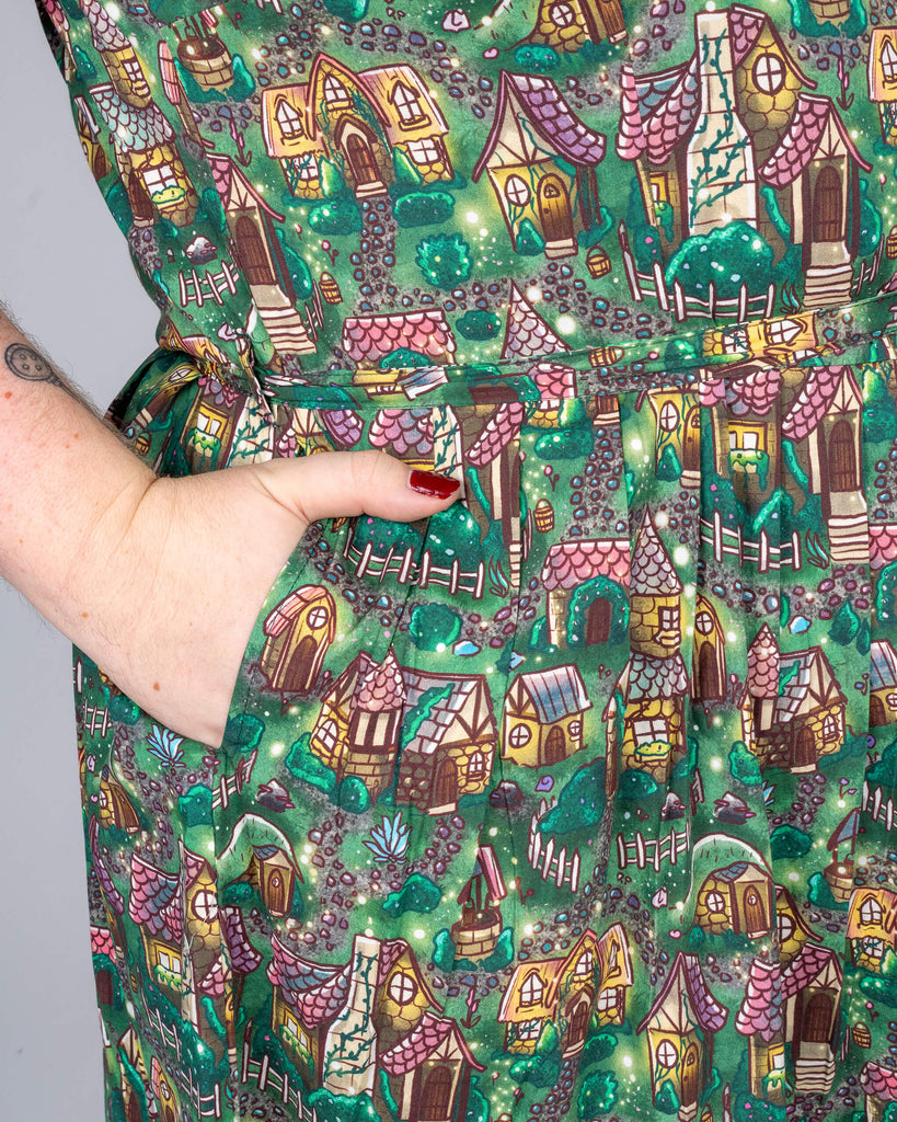 A detailed view of the cosy cottage print on Maddie’s dress, showing a hand in the pocket, which features various small houses, fences, and trees in rich forest greens and browns. The intricate design is clear, with Maddie’s red nail polish adding a pop of contrast.
