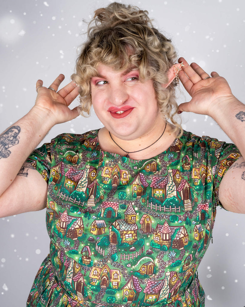  A close-up of Maddie’s facial expression and the intricate details on the cosy cottage dress, showing cartoon-like houses and forest elements in shades of green and brown. Maddie smiles mischievously, lifting both hands toward their pointed elf-like ears for a whimsical touch.