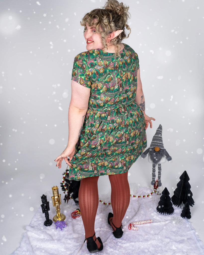  Maddie stands with their back to the camera, gently lifting the skirt of the cosy cottage dress, showcasing the dress’s vibrant pattern of cottages. Snow falls around them, and the wintery scene is set with festive decorations, including mini Christmas trees and holiday figurines.