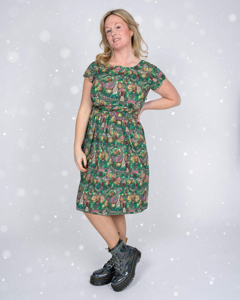 Karina poses side-on, showcasing the flattering fit of the “Christmas Cottages” dress, complete with a tie waist and detailed festive print.