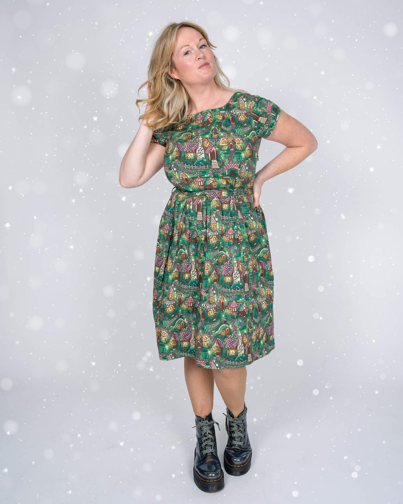 A full-length shot of Karina in the “Christmas Cottages” dress by Run & Fly, styled with iridescent boots for a fun, festive look. Snowfall effect adds a seasonal charm.