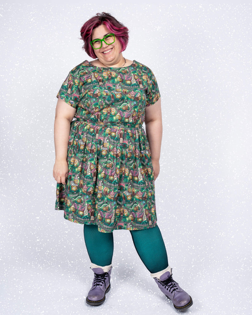 Jackie wears a Run & Fly tea dress featuring the “Cosy Cottage” print by Lauren Ash Design. The design showcases whimsical cottages, garden paths, and vibrant greenery on a rich green background, paired with teal leggings and lilac boots.