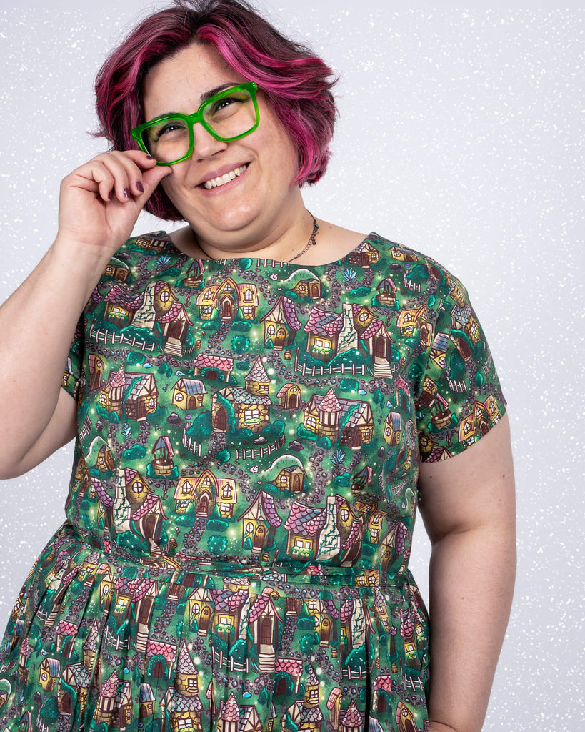 Close-up of Jackie smiling while adjusting green glasses. The “Cosy Cottage” print by Lauren Ash Design highlights charming cottages, rustic fences, and magical garden details on the Run & Fly 