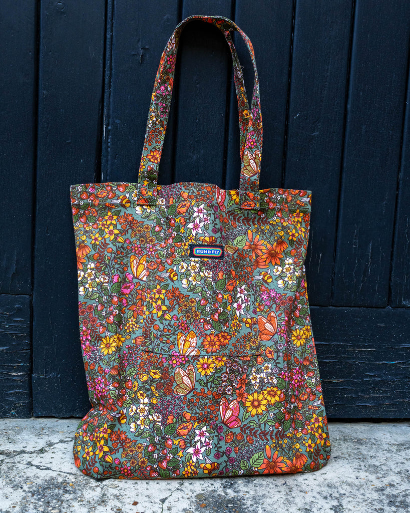 A close-up shot of a Run & Fly tote bag in the “Cottagecore” print by Jen James Design. The colourful floral pattern, with butterflies, flowers, and bees, stands out against the black wooden backdrop, with the Run & Fly logo patch visible on the front pocket.