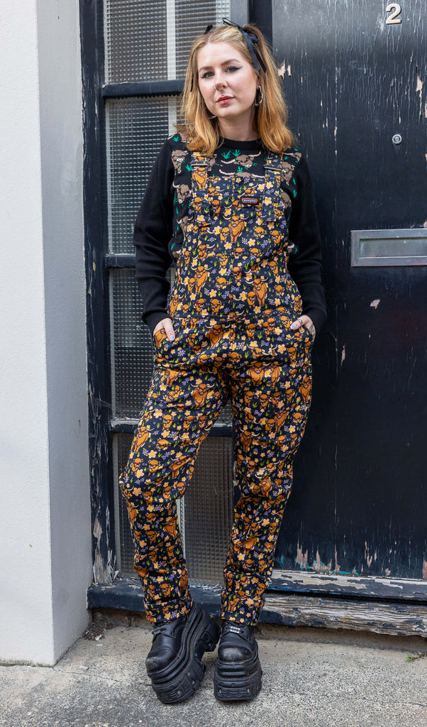 Florence, a femme model with blonde hair, is stood outside in Hove wearing Highland Cows Stretch Twill Dungarees with Highland Cow Jumper underneath and chunky black boots. The dungarees have an all over Highland Cow design. Florence is posing facing the camera with her hands in the dungaree front pockets. 