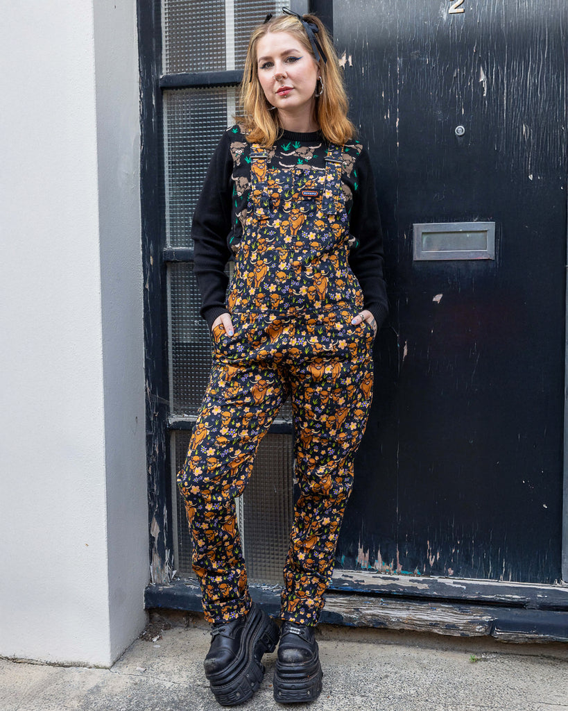 Florence, a femme model with blonde hair, is stood outside in Hove wearing Highland Cows Stretch Twill Dungarees with Highland Cow Jumper underneath and chunky black boots. The dungarees have an all over Highland Cow design. Florence is posing facing the camera with her hands in the dungaree front pockets. 
