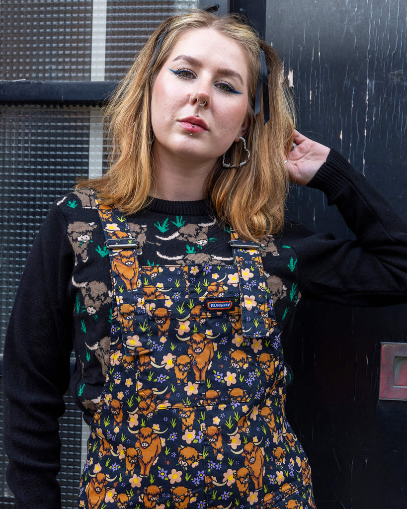 Florence, a femme model with blonde hair, is stood outside in Hove wearing Highland Cows Stretch Twill Dungarees with Highland Cow Jumper underneath. The dungarees have an all over Highland Cow design. Florence is posing toward the camera.