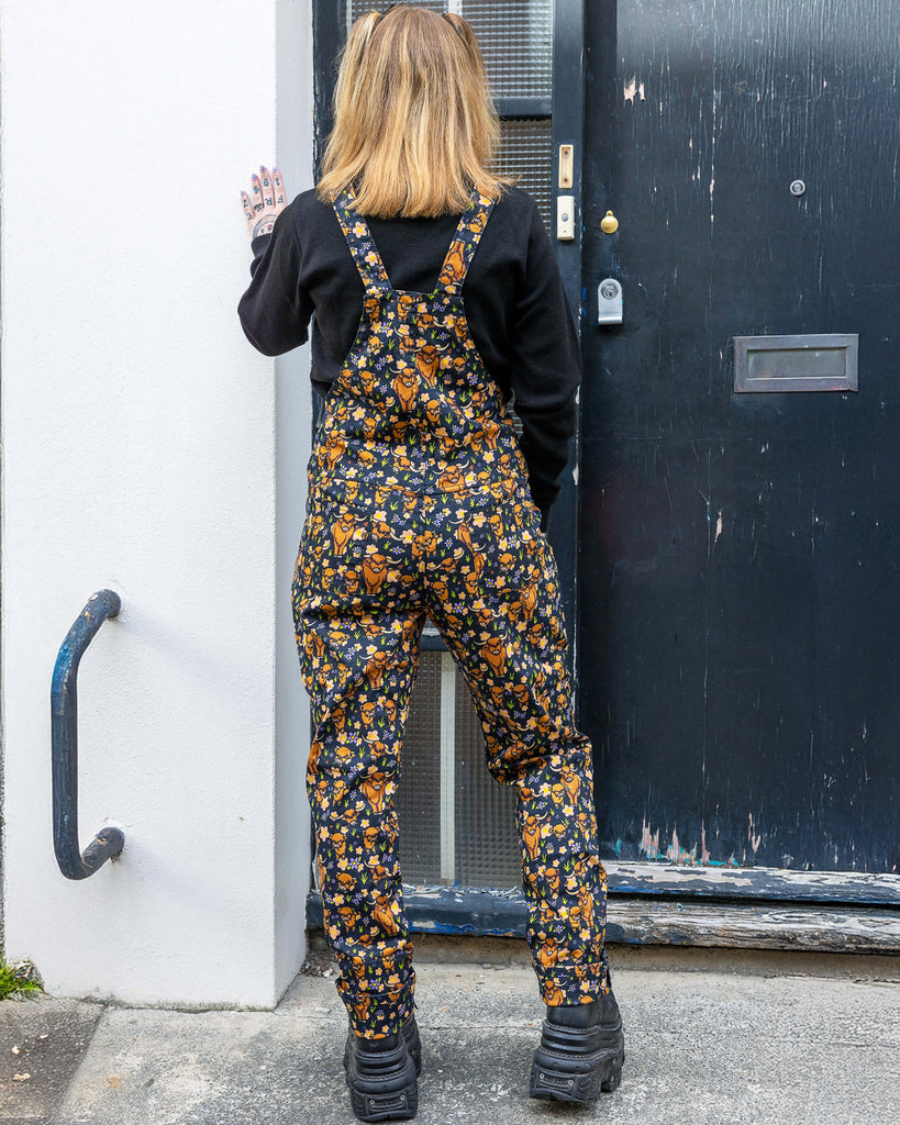 Florence, a femme model with blonde hair, is stood outside in Hove wearing Highland Cows Stretch Twill Dungarees with Highland Cow Jumper underneath and chunky black boots. The dungarees have an all over Highland Cow design. Florence is posing with her back to the camera and one hand leaning on a wall. 
