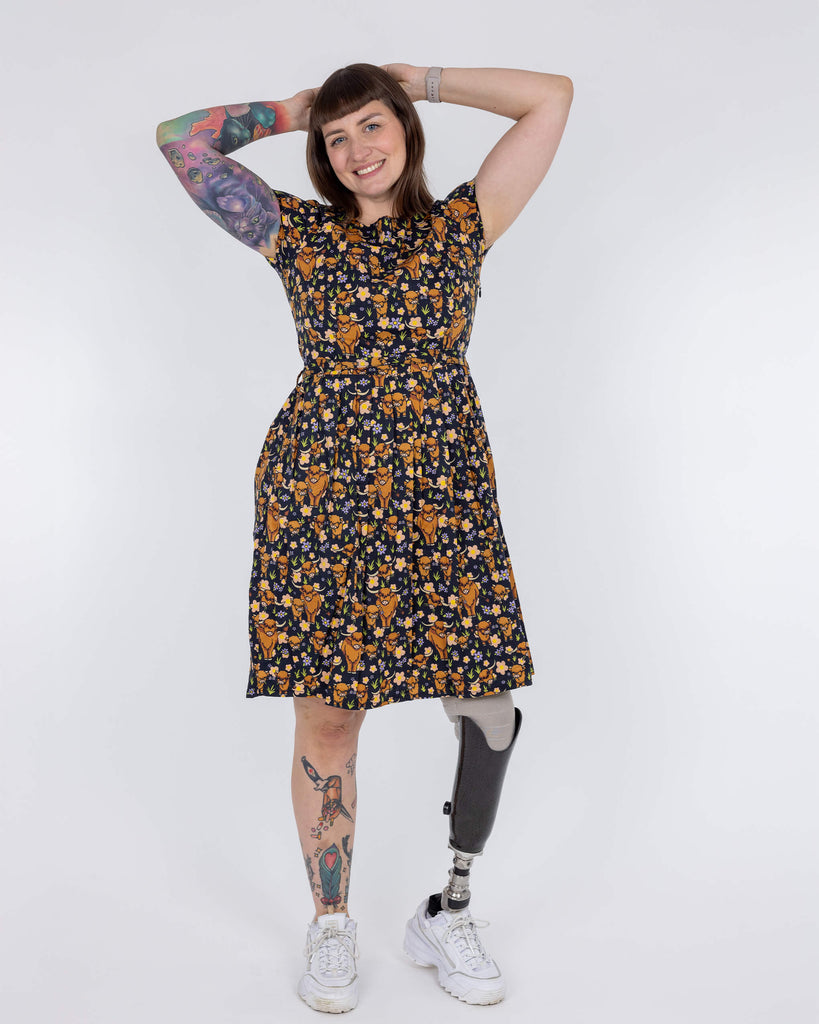 Laura is wearing a highland cow printed tea dress in front of a white background in a studio in Hove. She has a prosthetic leg and is smiling at camera with her hands above her head