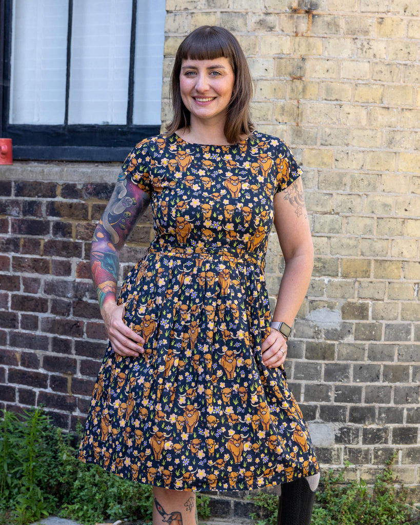 Laura is wearing a highland cow printed tea dress and is smiling at the camera with her hands resting on the dress. The photo is taken outside in a mews in Hove