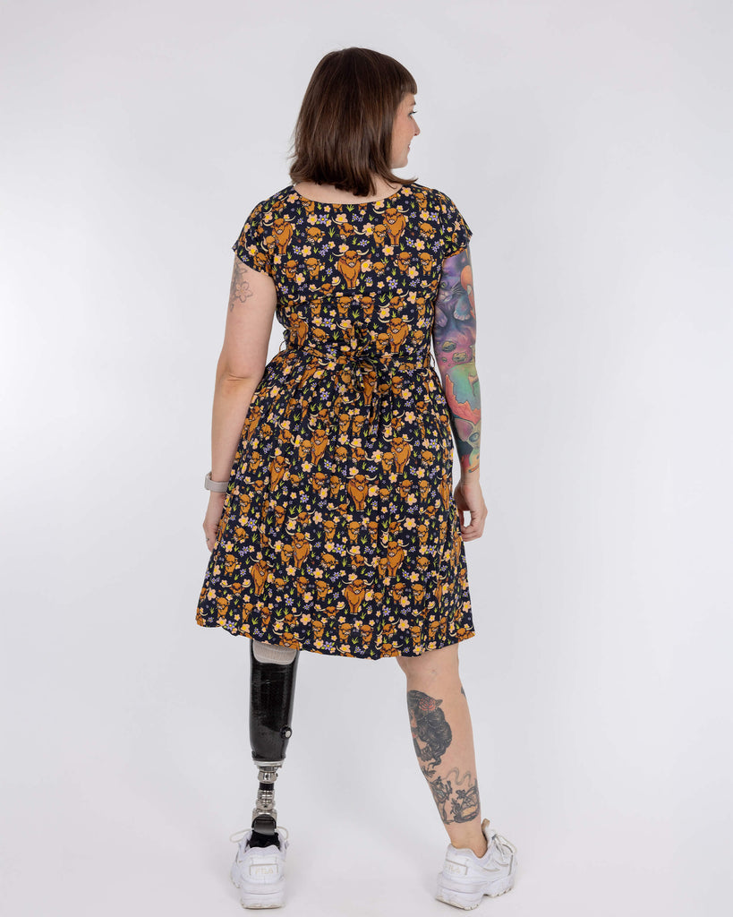 Laura is wearing a highland cow printed tea dress in front of a white background in a studio in Hove. She has a prosthetic leg and is posing with her back to the camera