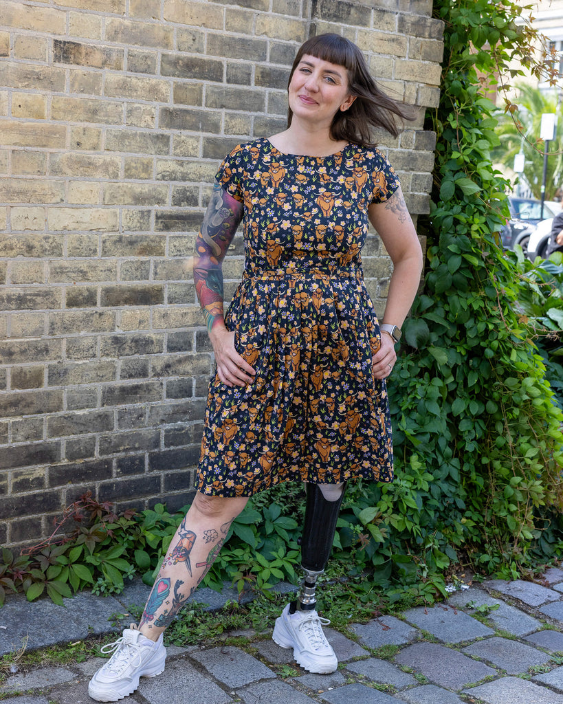 Laura is wearing a highland cow printed tea dress and her hair is blowing in the wind. She has a prosthetic leg and is smiling at camera
