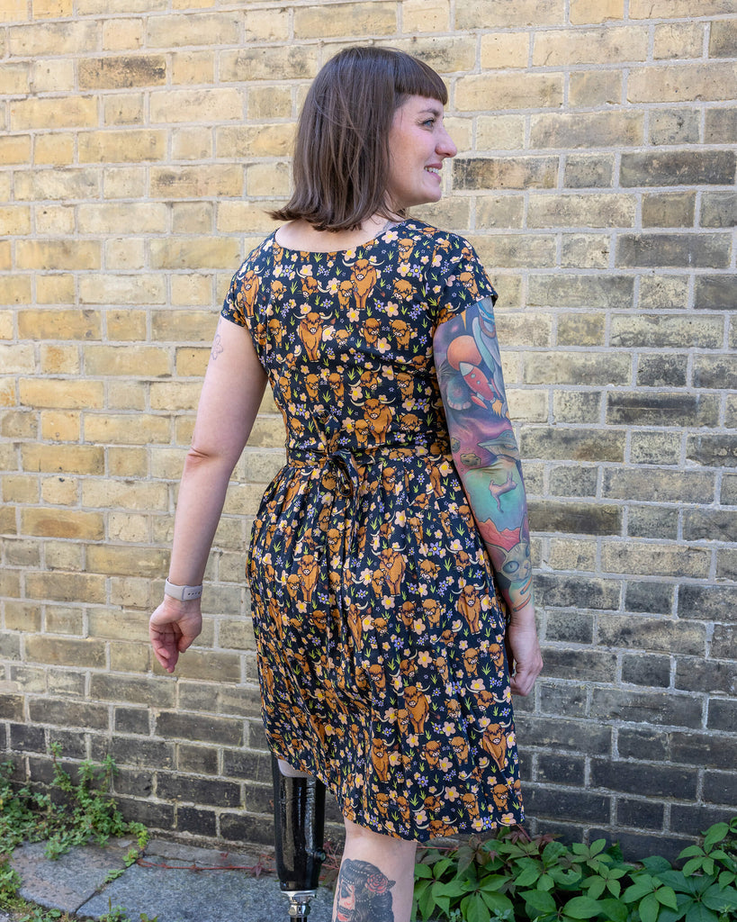 Laura is wearing a highland cow printed tea dress and her hair is blowing in the wind. She his stood with her back to the camera and is smiling looking off to one side 