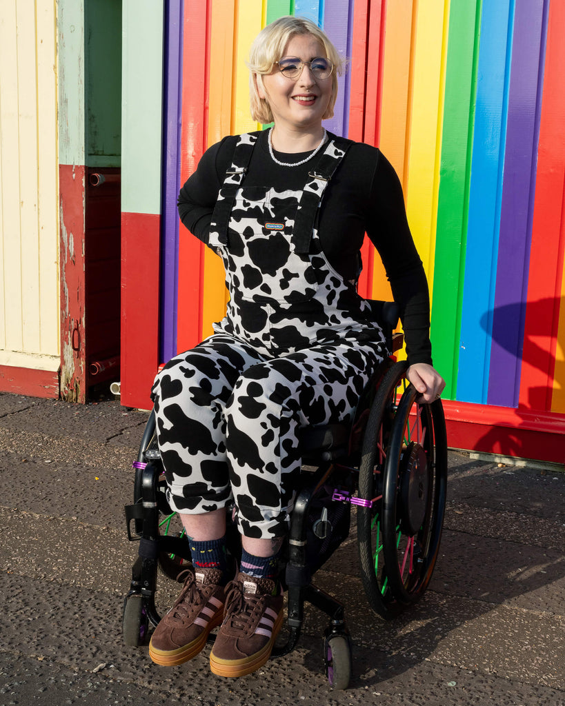 The model sits confidently in a wheelchair wearing Run & Fly Cow Print Stretch Twill Dungarees, posing in front of a rainbow wall. The black and white cow print adds a playful and unique touch, exemplifying the ethical slow fashion brand’s quirky style.