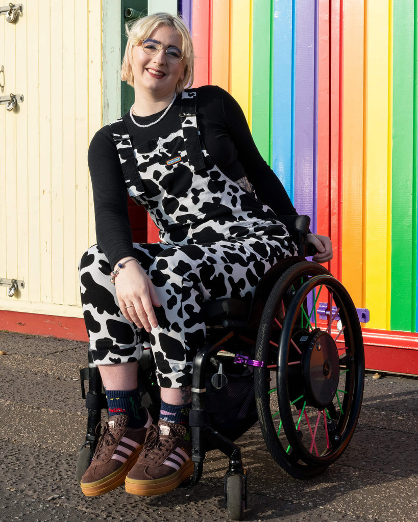 The model leans sideways, smiling in Run & Fly Cow Print Stretch Twill Dungarees with a black top. The dungarees, by an ethical slow fashion brand, showcase a fun cow print pattern, adding a unique twist to this casual, stylish outfit.