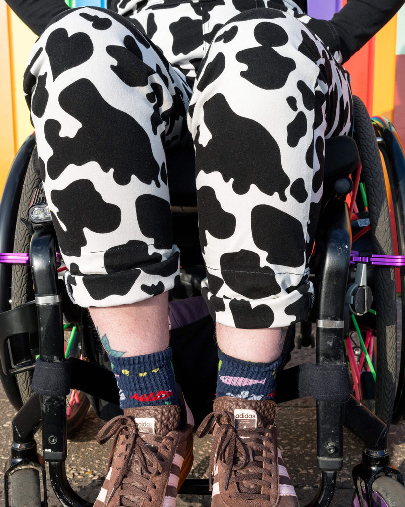 Close-up of the model’s legs in Run & Fly Cow Print Stretch Twill Dungarees, paired with brown trainers and fun socks. The ethical slow fashion brand's dungarees feature a distinctive cow print, adding a quirky style to this comfortable look.
