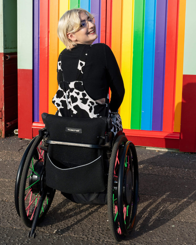 The model faces away from the camera, showing the back of the Run & Fly Cow Print Stretch Twill Dungarees. The colourful rainbow wall adds a bright, joyful contrast to the black and white cow print design by this ethical slow fashion brand.