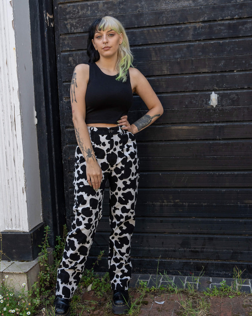 Model wearing Run & Fly’s cow print jeans, posing with one hand on the hip in front of a rustic door. The high-waisted, straight-leg jeans are paired with a black crop top, highlighting the playful and quirky cow print design. Ethically produced by a small slow fashion company.