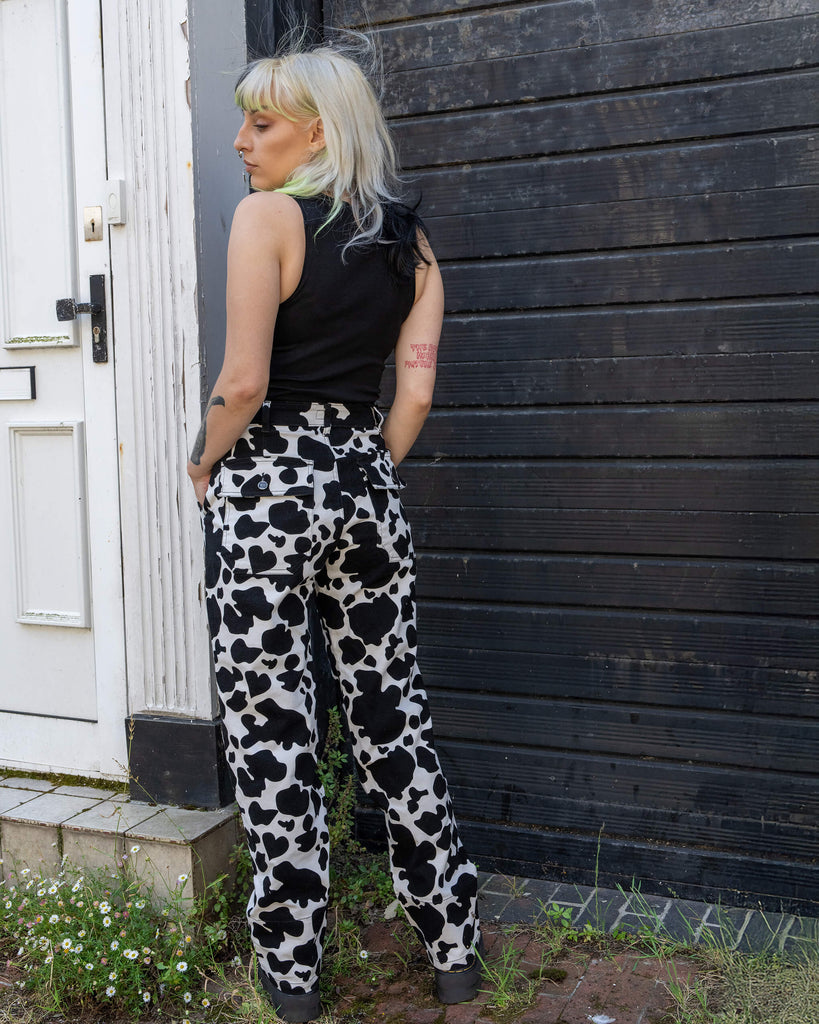 Model showing the back view of Run & Fly’s cow print jeans, standing against a dark wood backdrop. The high-waisted design features two back pockets and a relaxed, straight-leg fit. A playful and quirky print from an ethical, small fashion brand, perfect for a bold, sustainable style.
