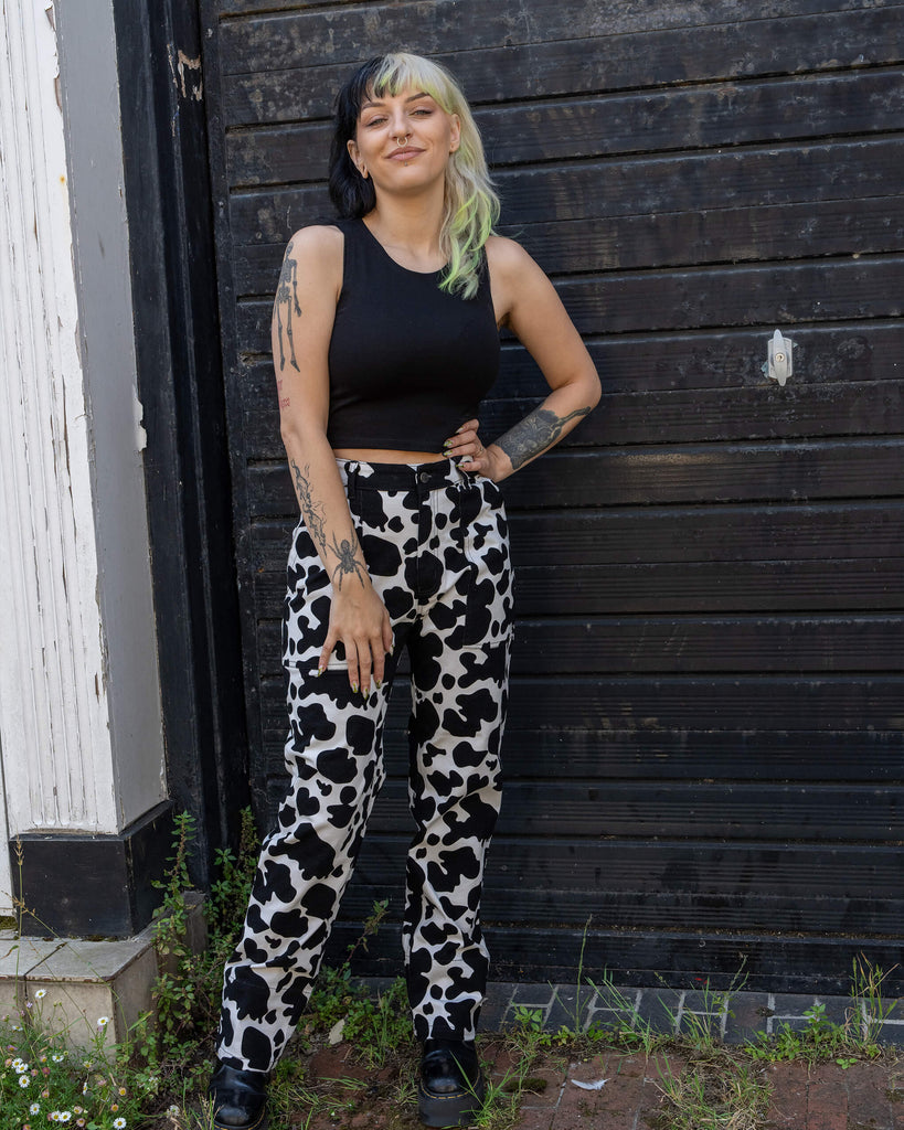 Model wearing black and white cow print jeans from Run & Fly, a small ethical slow fashion brand. The model stands in front of a dark, rustic door, with hands on hips, showing off the high-waisted, straight-leg fit. The bold cow print creates a fun, standout look paired with a simple black crop top. The jeans offer a statement style with ethical, slow fashion production.