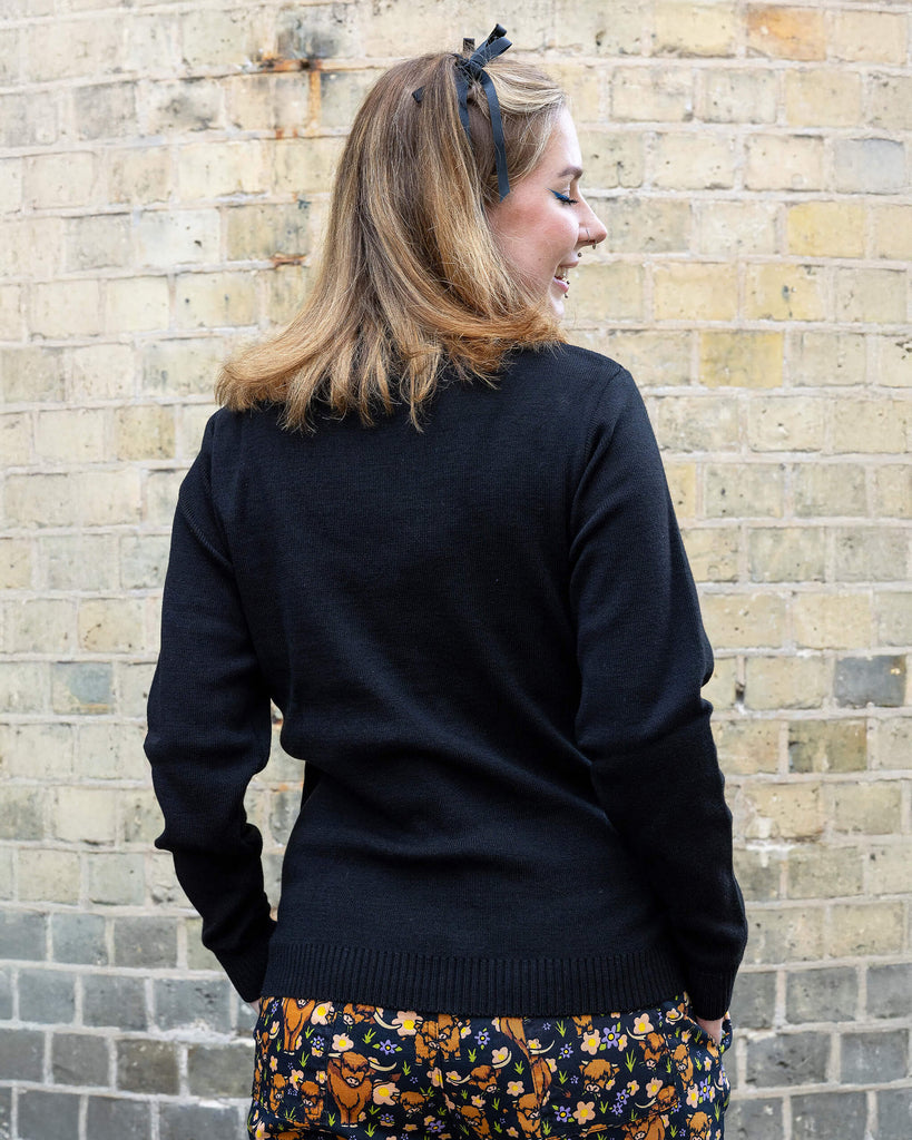 Florence, a femme model with blonde hair is stood outside in Hove wearing Highland Cow Jumper on top of Highland Cow Dungarees. The jumper is black with highland cows on the torso. Florence is stood with her back to the camera to show plain black back of jumper.