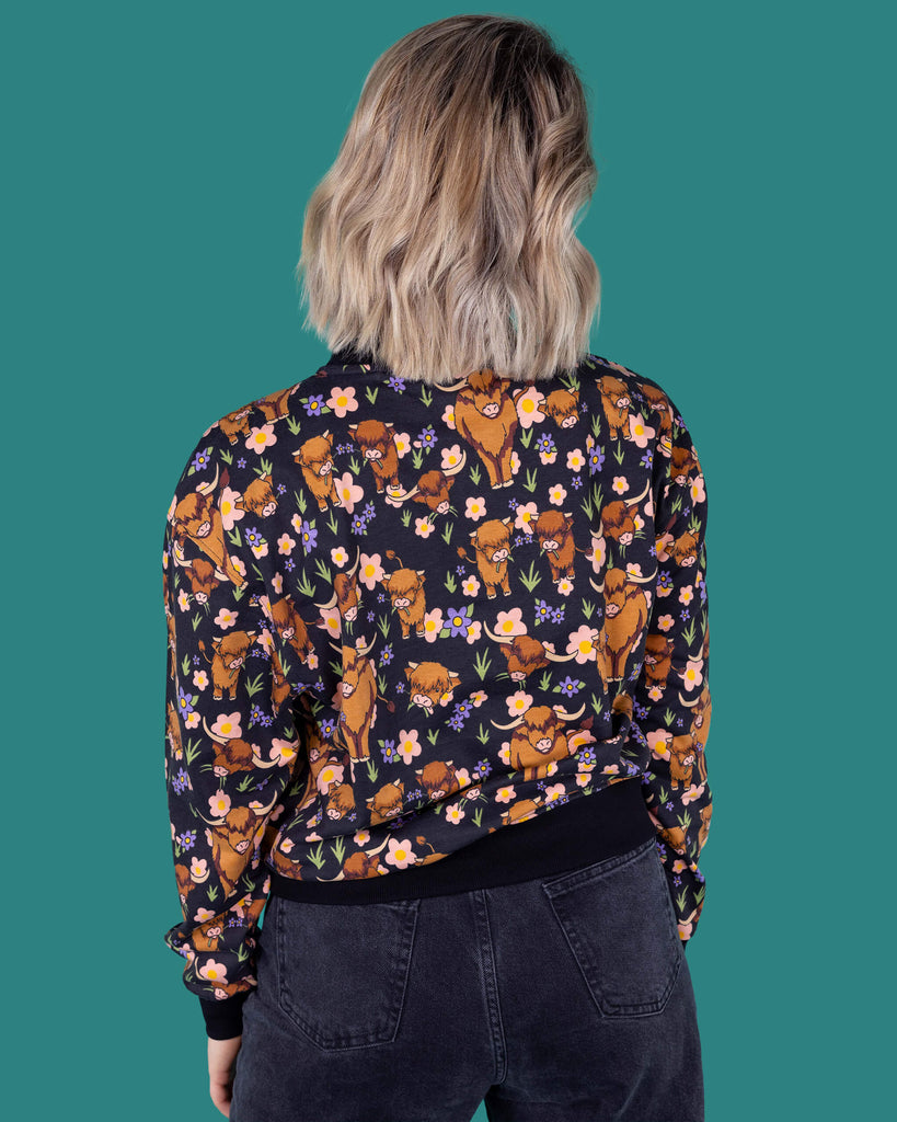 The Highland Cow Sweatshirt worn by a femme model with short blonde hair with dark denim jeans on a teal studio background. She is facing away from camera with both hands resting by her side. The black base sweatshirt has pink, yellow and purple flowers, grass bunches and various brown highland cows all over with a black neck, hem and sleeve trim.
