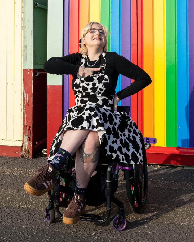 Eliza poses with a smile in Run & Fly’s cow print flared pinafore dress, sitting in front of a rainbow-coloured wall. The dress's black and white cow print and playful flare skirt are styled over a black top with brown trainers. Ethical slow fashion by Run & Fly.