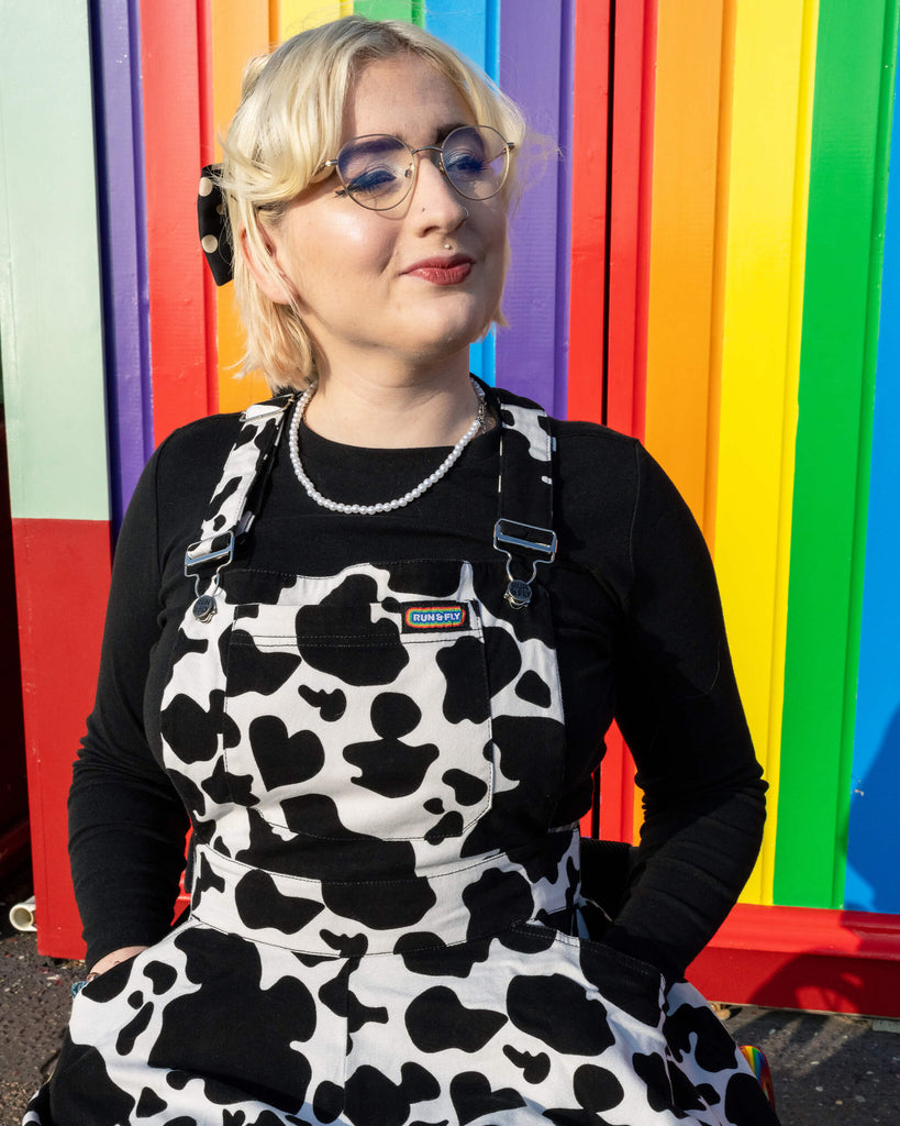 Close-up of Eliza wearing Run & Fly’s cow print flared pinafore dress, featuring a black long-sleeve top underneath and a pearl necklace. The dress's black and white cow print contrasts with the rainbow wall in the background, emphasising Run & Fly’s ethical, inclusive, slow fashion approach.