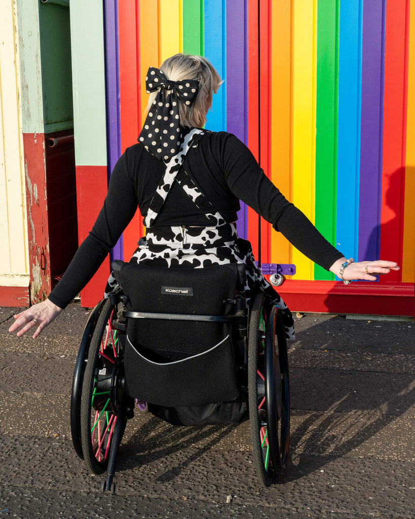 Back view of Eliza wearing Run & Fly’s cow print flared pinafore dress, with a polka dot hair bow and the vibrant rainbow wall as a backdrop. The adjustable straps of the dress cross over, adding a cute detail to this ethical, slow fashion piece by Run & Fly.