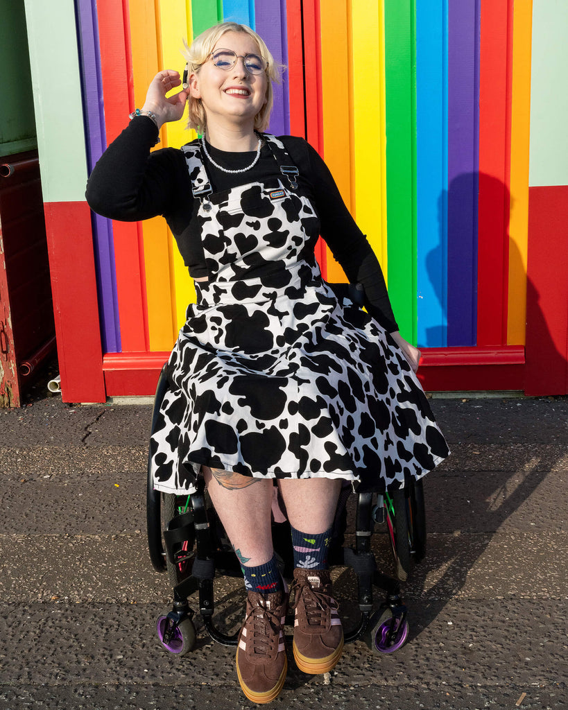 Eliza models Run & Fly’s cow print flared pinafore dress, sitting in a wheelchair with a bright rainbow wall behind. The playful black and white cow pattern pops against the colourful background. The dress is layered over a black top with brown trainers and rainbow socks, showcasing Run & Fly's commitment to inclusive, ethical slow fashion.