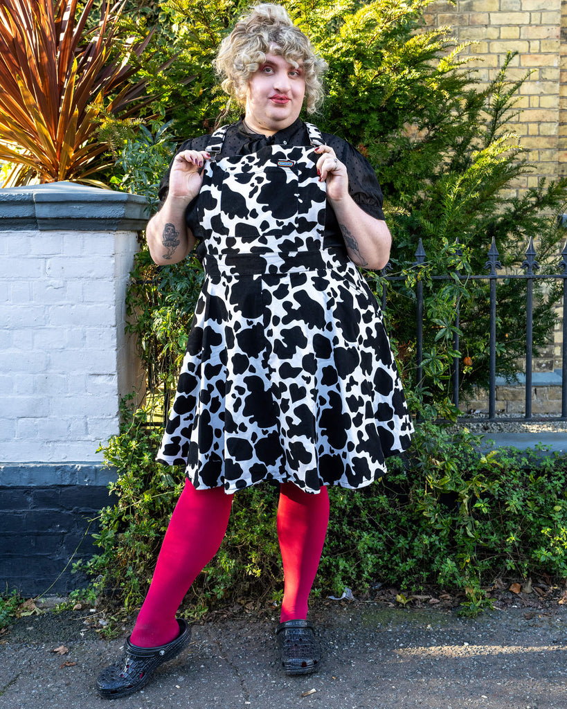 Maddie Twirling in Cow Print Pinafore - Maddie strikes a playful pose in the Run & Fly cow print pinafore, holding out the skirt to show its flared style. She pairs the dress with bright pink tights and black footwear, standing against a garden setting with greenery.