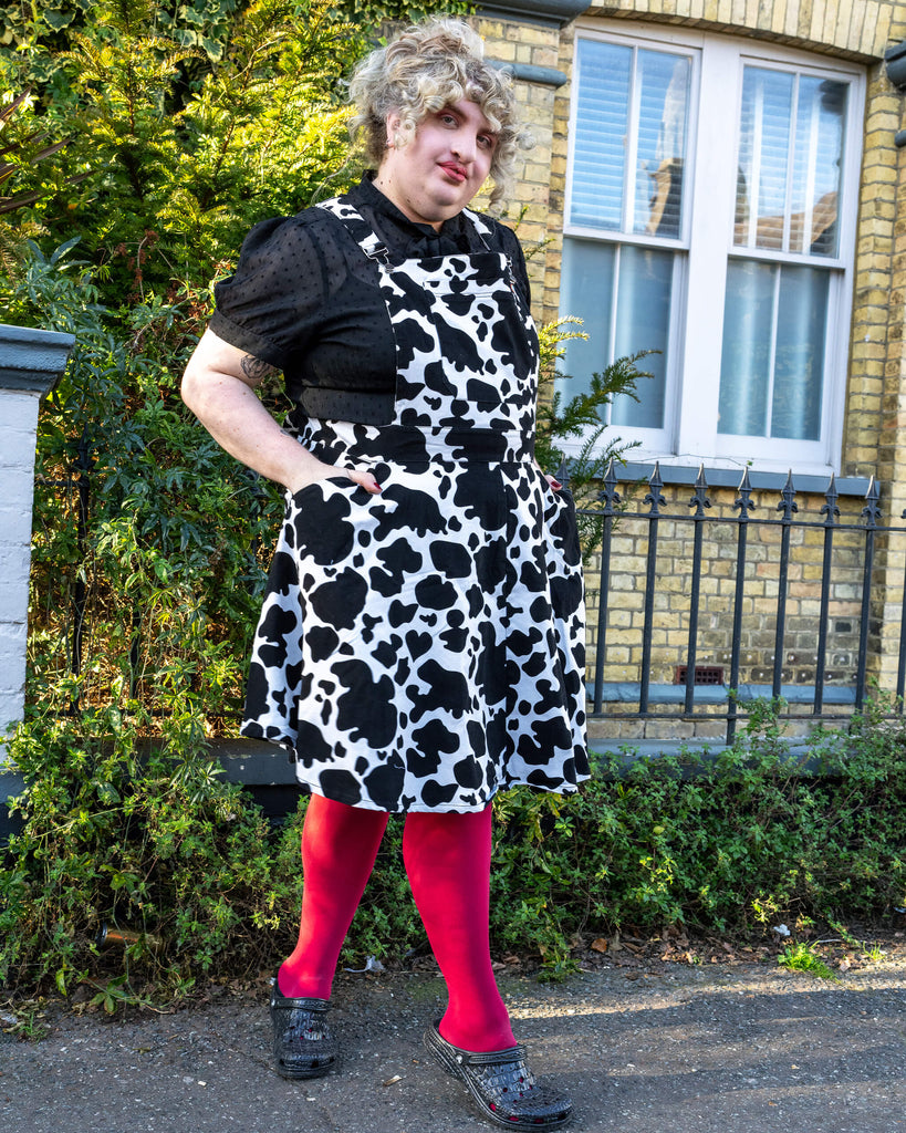 Maddie Posing in Run & Fly Cow Print Pinafore - Maddie stands outdoors, wearing the Run & Fly black and white cow print pinafore paired with bright pink tights and black shoes. The outfit is styled with a black textured blouse, adding a playful pop of colour against a green leafy background.