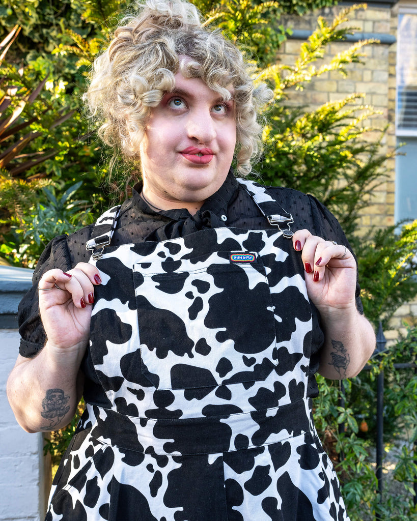 Playful Pose in Run & Fly Cow Print Pinafore - Maddie poses with her hands near the straps of the pinafore, showing off the vibrant cow print pattern. The black blouse and pink tights add to the playful, unique style of the outfit.
