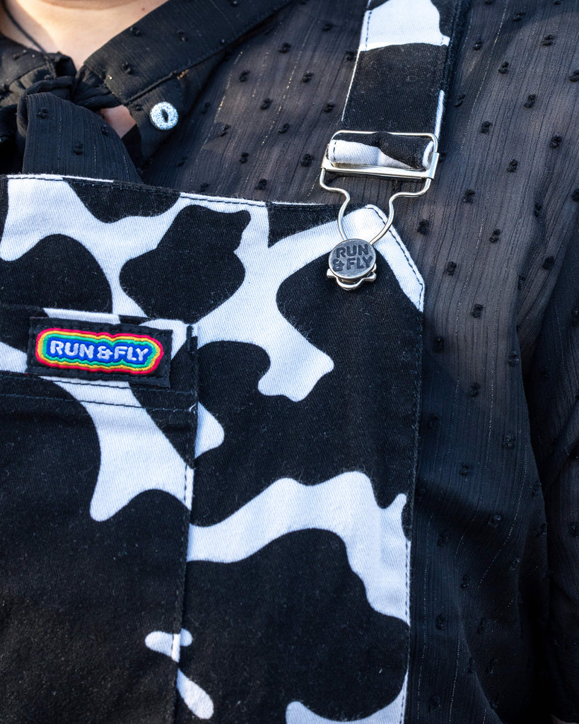 Close-up of Maddie’s Run & Fly Cow Print Pinafore - A close-up view of the Run & Fly logo badge on a black and white cow print pinafore dress, featuring a vibrant rainbow tag. Maddie wears a textured black blouse underneath.