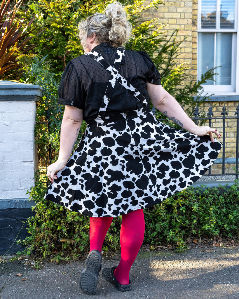Back View of the Cow Print Pinafore - Maddie shows the back of the cow print pinafore, with crossed straps over a black blouse. The skirt flares out, creating a classic pinafore silhouette. Bright pink tights add a bold contrast to the black and white print.