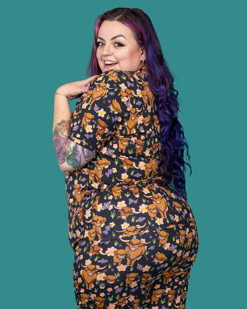 The Highland Cows Short Sleeve T Shirt worn by a femme alternative tattooed model with long pink and purple hair, the matching cycle shorts on a teal studio background. She is facing away laughing looking over her left shoulder with her hand resting on her shoulder. The black base t shirt features grass, purple and yellow flowers and brown highland cows eating grass.