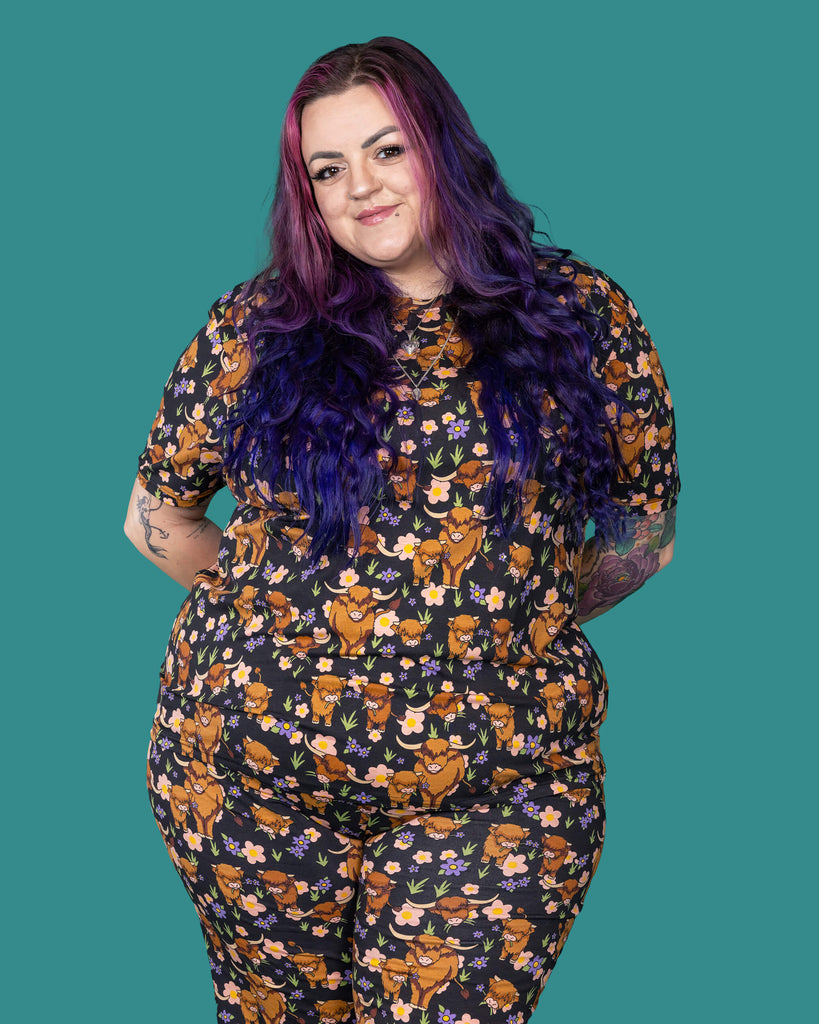 The Highland Cows Short Sleeve T Shirt worn by a femme alternative tattooed model with long pink and purple hair, the matching cycle shorts on a teal studio background. She is facing away laughing looking over her left shoulder with her hand resting on her shoulder. The black base t shirt features grass, purple and yellow flowers and brown highland cows eating grass.