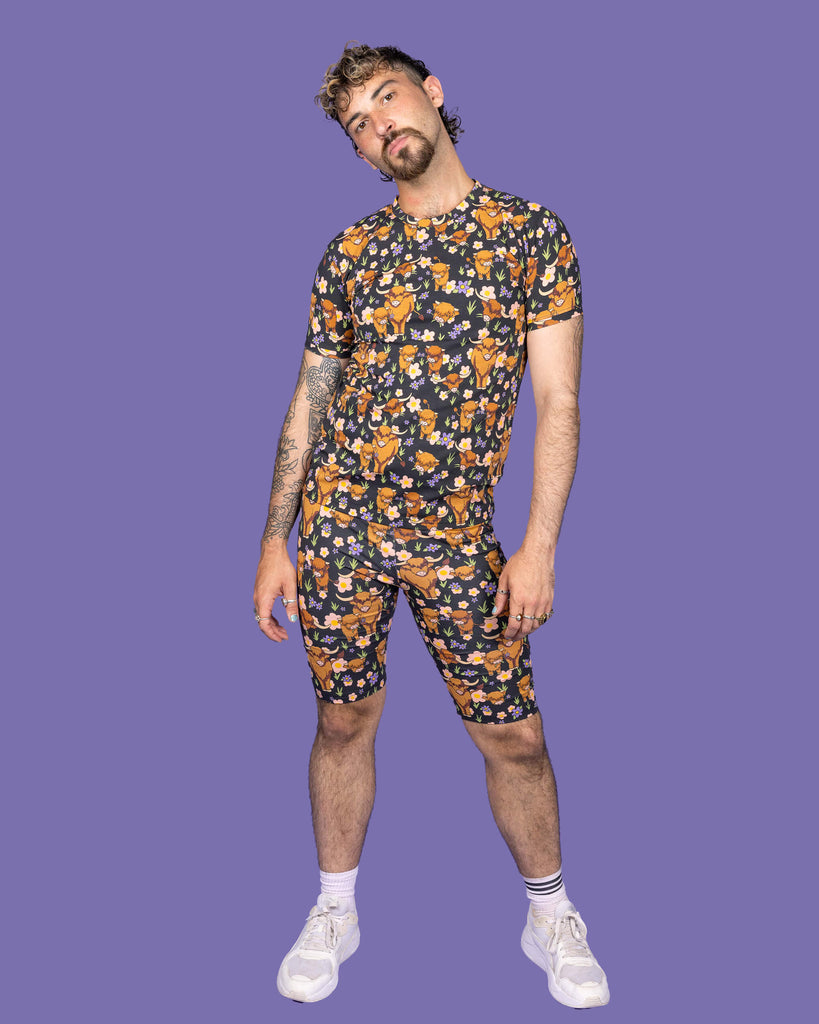 The Highland Cows Short Sleeve T Shirt worn by a masc non binary person with a short brown curly mullet with the matching cycle shorts and white trainers on a purple studio background. They are facing forward leaning more on their left hip and their head tilted left pouting to camera. The black base t shirt features grass, purple and yellow flowers and brown highland cows eating grass.