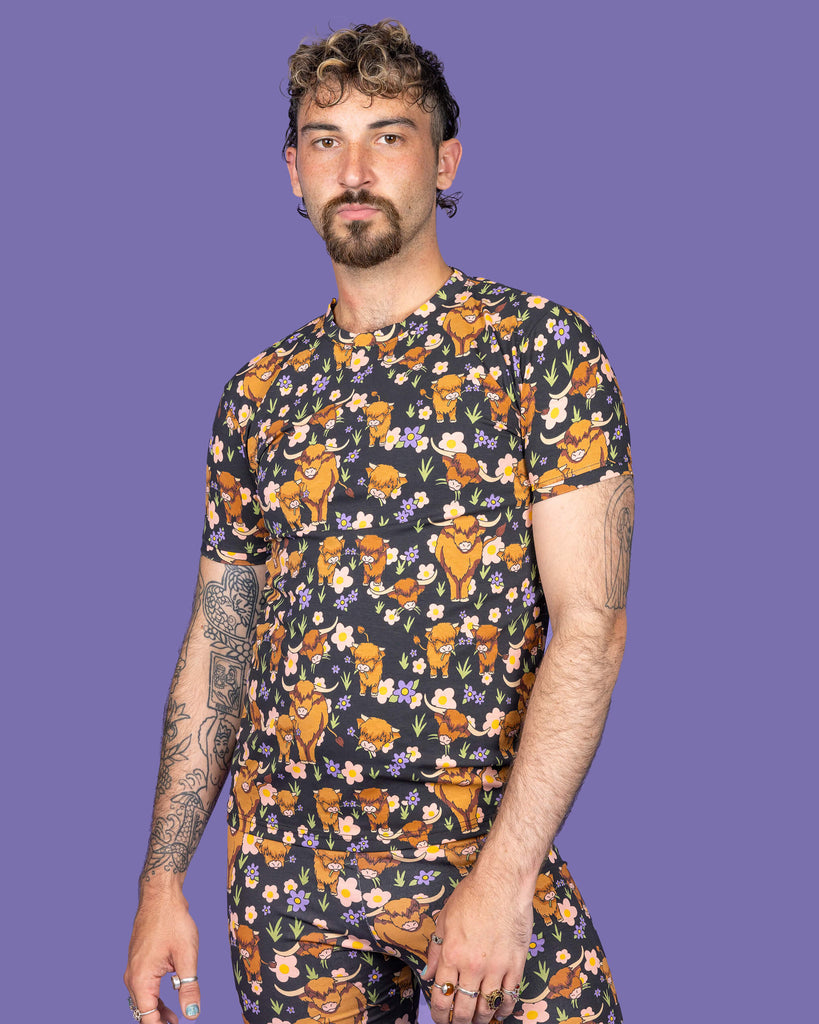 The Highland Cows Short Sleeve T Shirt worn by a masc non binary person with a short brown curly mullet with the matching cycle shorts on a purple studio background. They are facing forward pouting to camera leaning more on their left hip with their arms resting by their sides. The black base t shirt features grass, purple and yellow flowers and brown highland cows eating grass.