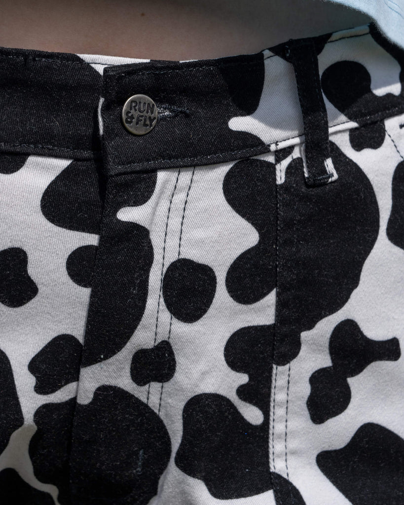A detailed shot of the button and waistband of Run & Fly's Black & White Cow Print Straight Leg Trousers. The metallic button reads "Run & Fly," while the cow print pattern in black and white contrasts on the cotton fabric. Ethical slow fashion brand Run & Fly focuses on fun, quirky designs with a retro aesthetic. The trousers are tailored for a comfortable, stylish fit.